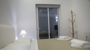 A hotel room with a bed, a nightstand with a lamp, a wooden coat rack, and a sliding door to a bathroom. - Villa Athena (Mykonos)