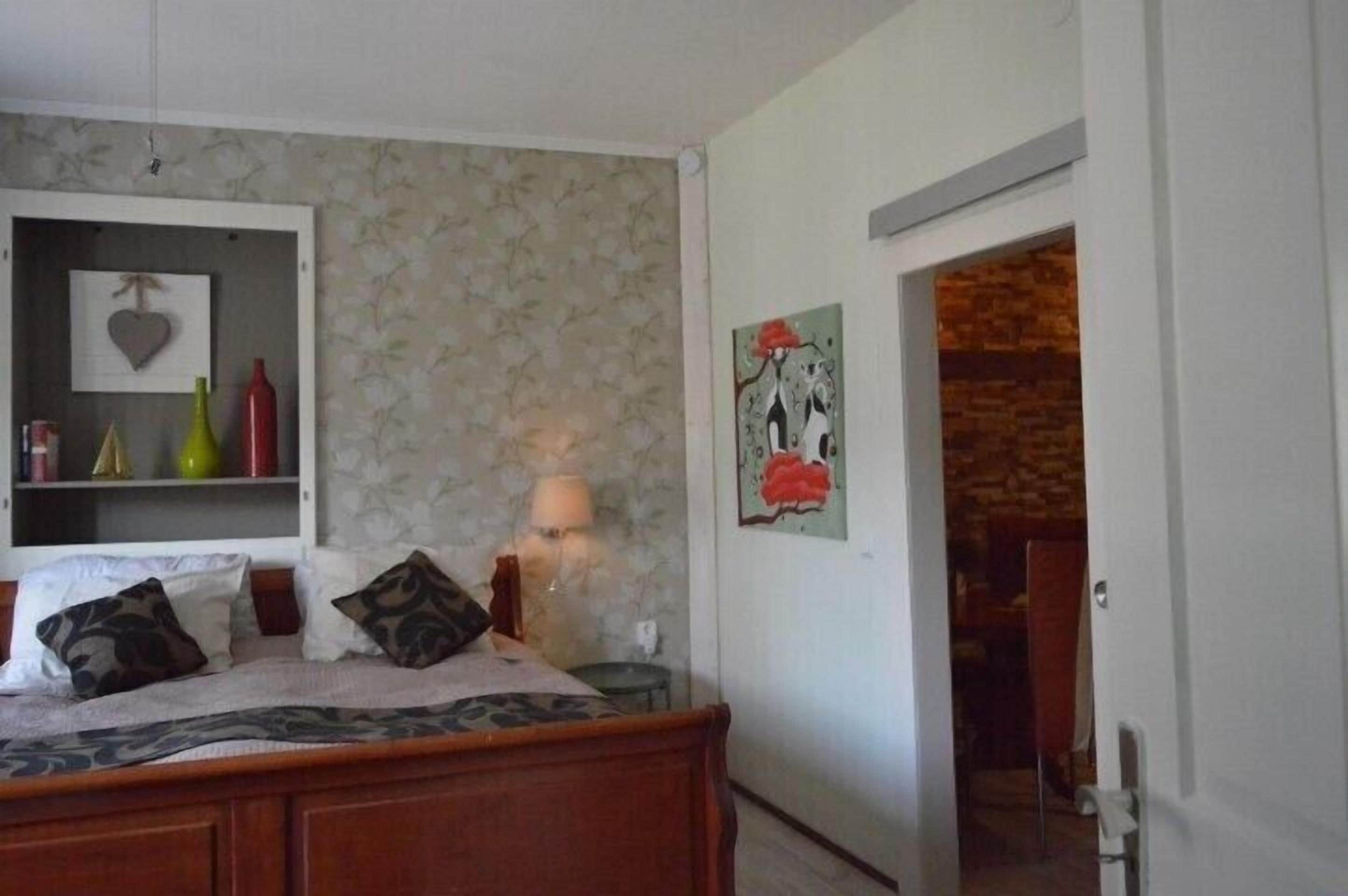 Un dormitorio con una cama, una mesita de noche con lámpara, un estante con adornos y una puerta que da a otra habitación.