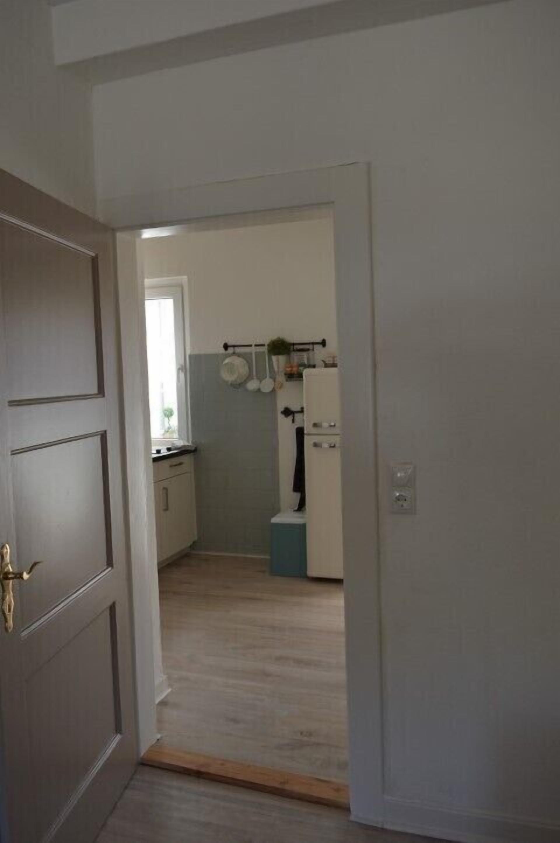 A narrow hallway leading to a kitchen with a refrigerator, a window, and a door with a brass handle.