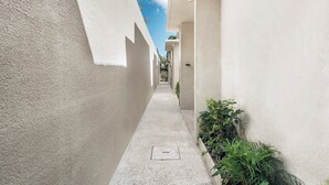 A narrow corridor with a concrete floor, white walls, and green plants on one side.