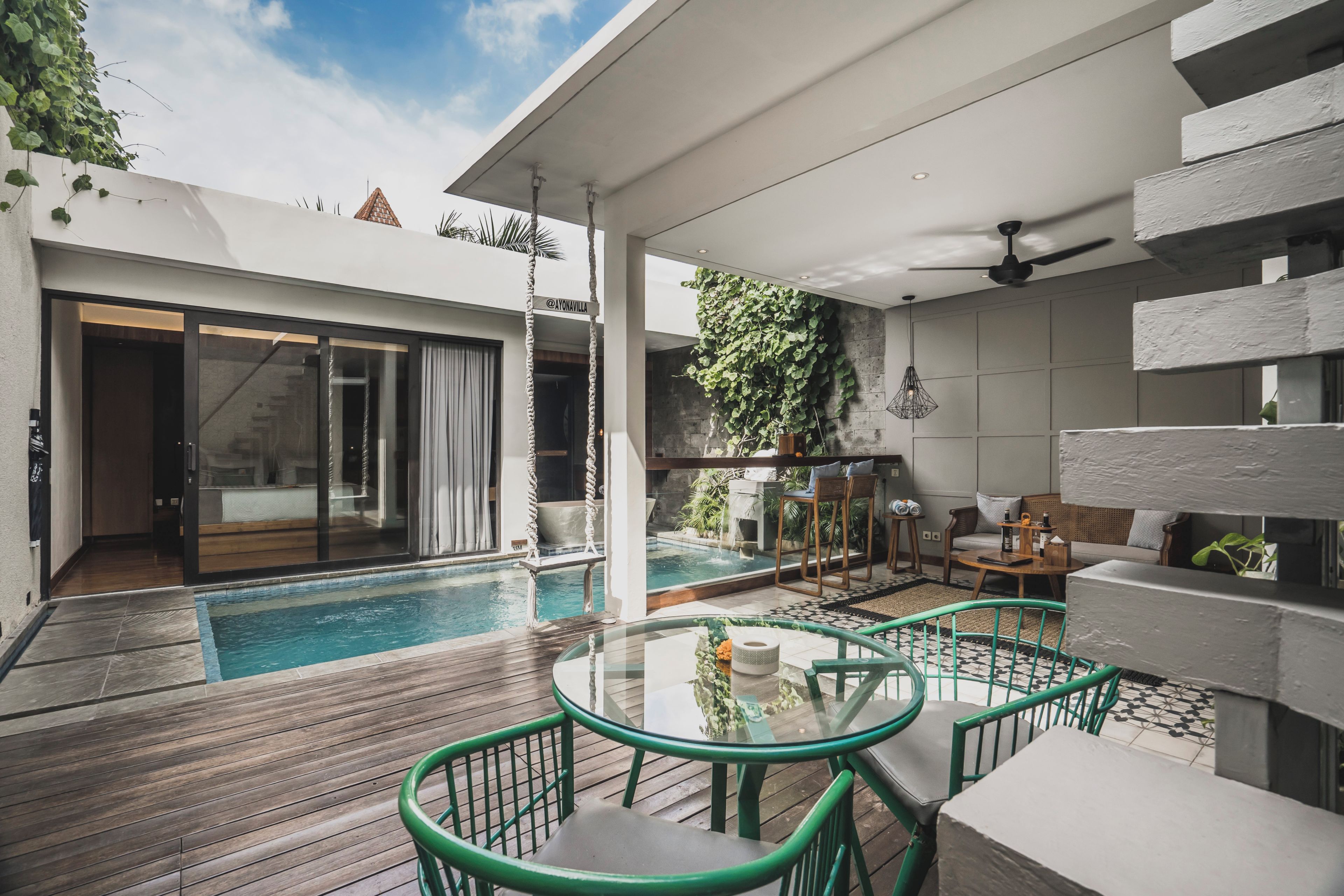 A modern outdoor pool area with a glass table and chairs, a seating area with a sofa and coffee table, and a view of a building with a large glass door.