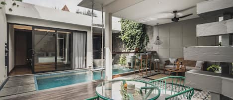 Un área de piscina moderna al aire libre con mesa y sillas de vidrio, una zona de estar con sofá y mesa de centro, y vista a un edificio con un gran ventanal.