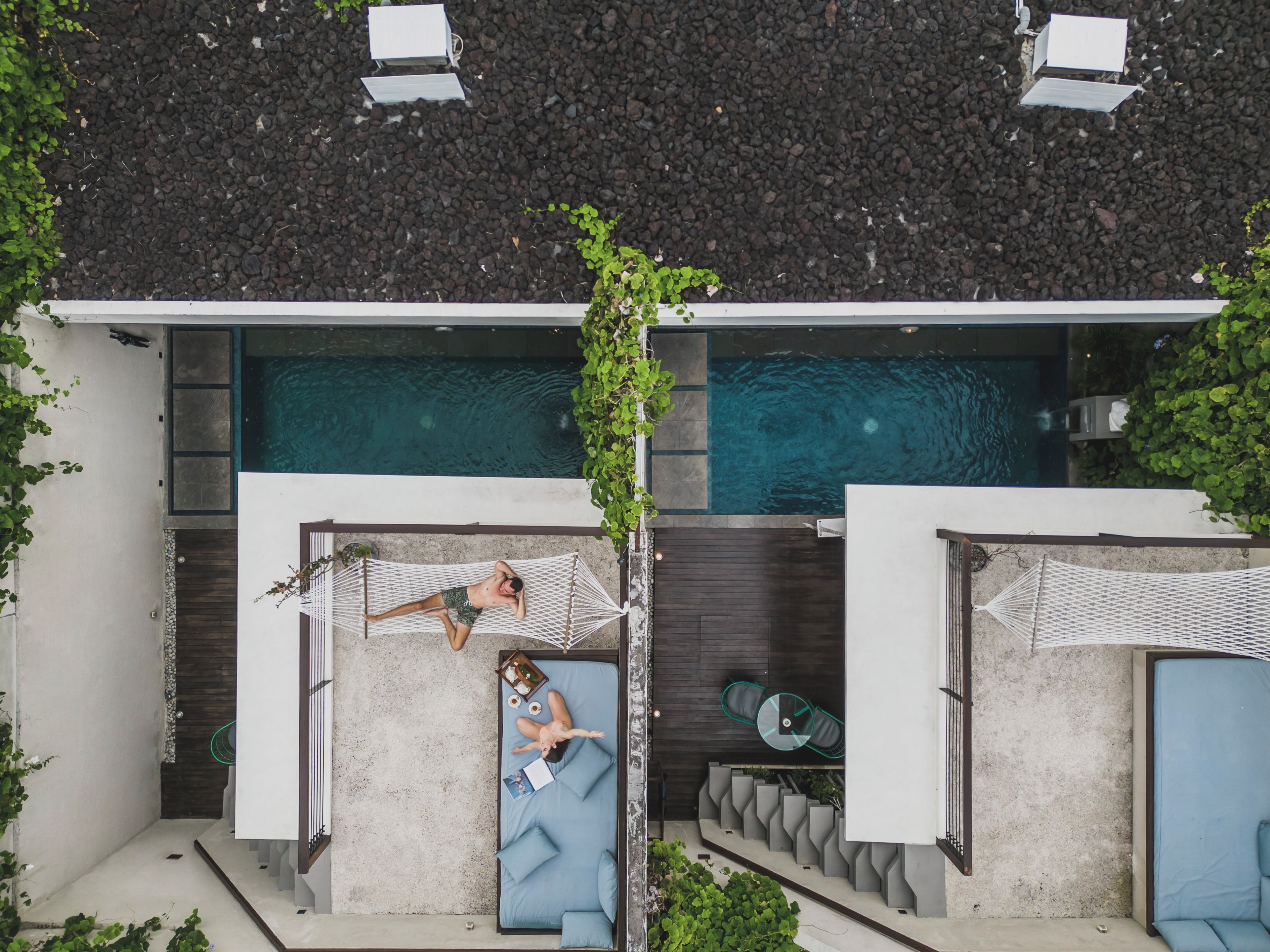 A modern outdoor pool area with two hammocks and a lounge chair, surrounded by greenery.
