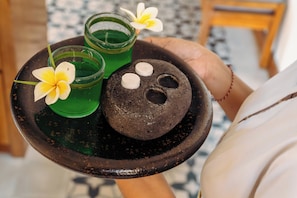 Une personne tenant un plateau sur lequel se trouvent une pierre, deux verres contenant un liquide vert et une fleur.