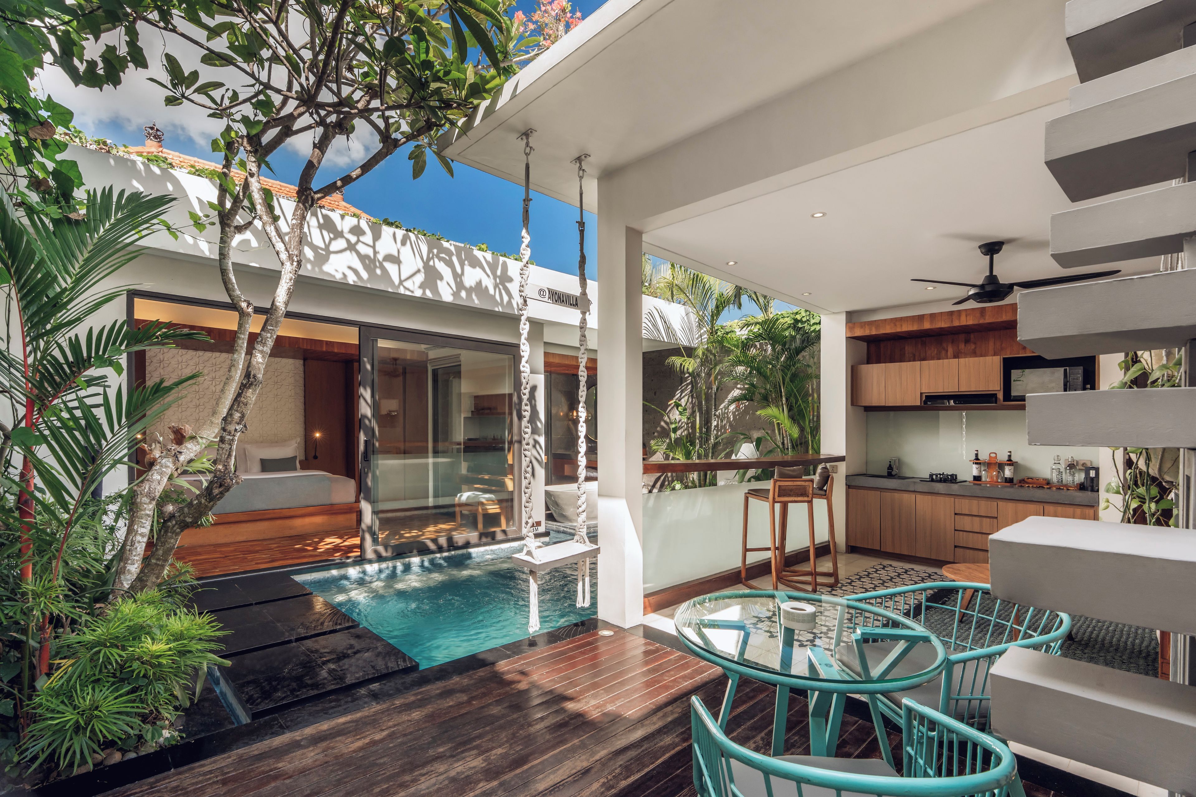 A modern outdoor pool area with a glass balustrade, wooden deck, and a dining area with a glass table and turquoise chairs.