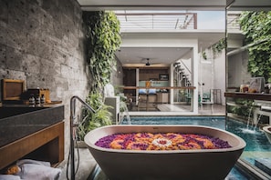 A modern bathroom with a freestanding bathtub filled with colorful flower petals, a stone wall, and a view of a pool area. - Ayona Villa Seminyak by Ini Vie Hospitality (Kerobokan)