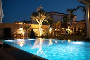 A swimming pool with a stone wall and palm trees, surrounded by outdoor seating and a thatched-roof structure. - Kaipo Alacati (Cesme)
