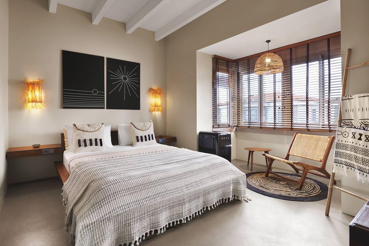 Un dormitorio con una cama grande, dos luces fijadas a la pared, un ventanal con persianas, una silla de ratán y una obra de arte en la pared.