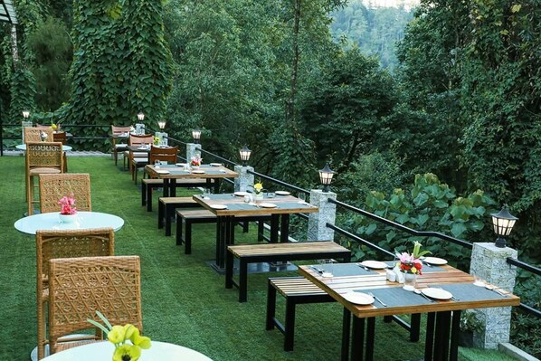 An outdoor dining area with multiple tables and chairs, surrounded by lush greenery and stone lanterns.