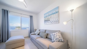 A modern living room with a sofa, ottoman, and a painting of a beach scene. - Refurbished 1-bed in popular Los Gigantes w/large terrace, sea & cliff views (Los Gigantes)