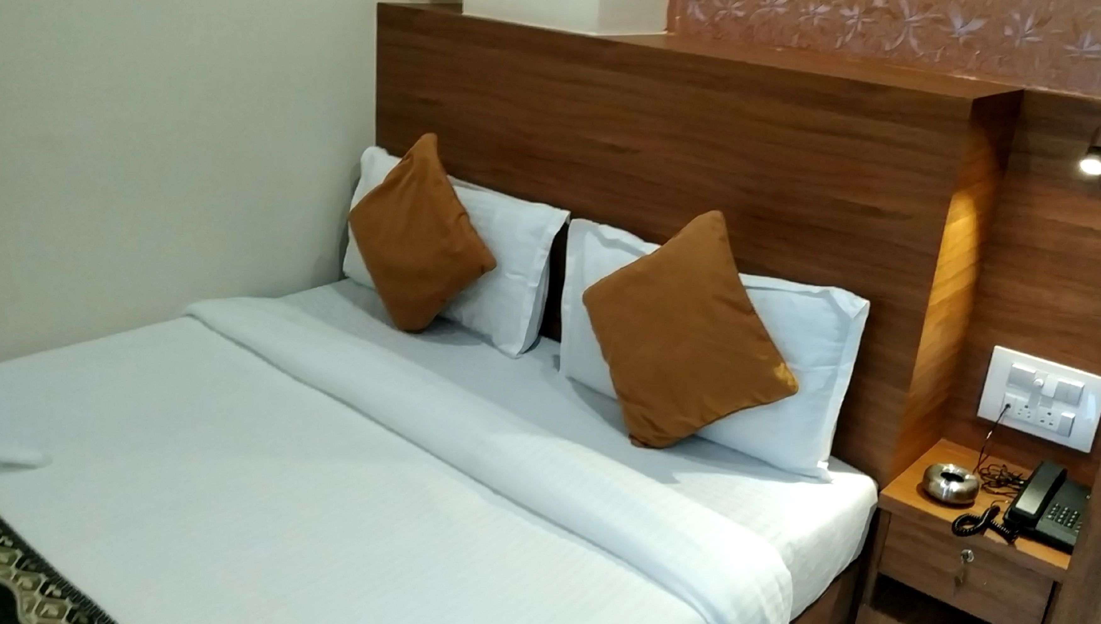 Une chambre d’hôtel avec une tête de lit en bois, un lit recouvert de draps blancs, deux oreillers marron, une table de chevet avec un téléphone et un chargeur, et une prise électrique au mur.