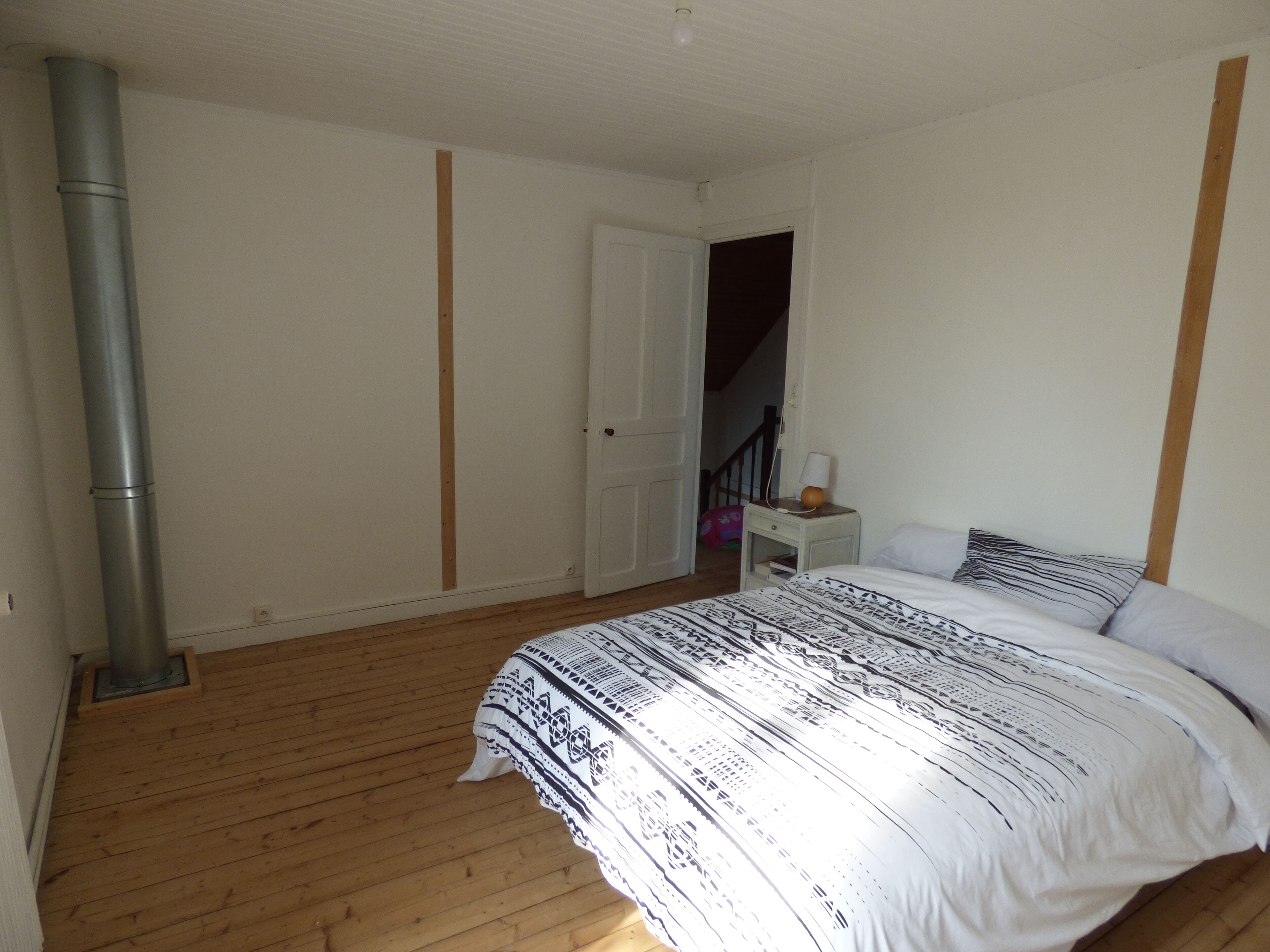 Ein Schlafzimmer mit Holzboden, einem Bett mit gemusterten Betttextilien, einem Nachttisch mit Lampe und einer weißen Tür, die in einen weiteren Raum führt.