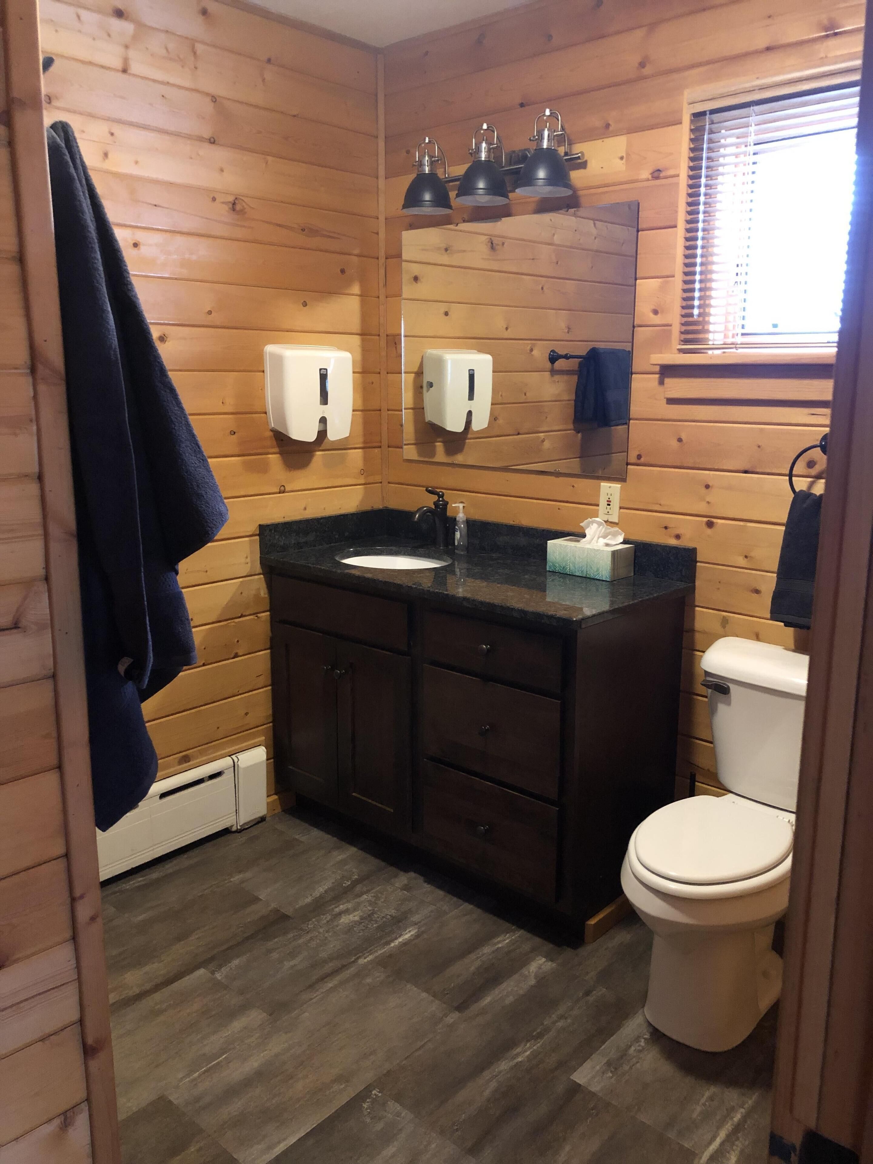 A wooden-paneled bathroom with a sink, mirror, three wall-mounted lights, a toilet, and a towel rack.