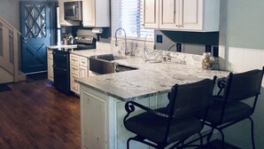 Cocina con gabinetes blancos, un lavamanos de acero inoxidable y una encimera de mármol. Se observa una silla y una estufa, ambos de color negro.