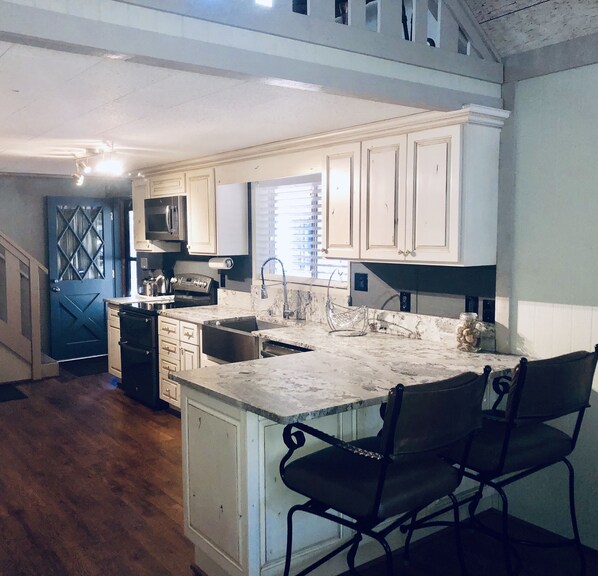 A kitchen with white cabinets, a stainless steel sink, and a marble countertop. There is a black chair and a black stove. - Modern cozy Forrest Lakes Cabin (Forrest Lake)
