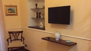 A hotel room with a bed, a chair, a flat-screen TV mounted on the wall, a shelf with decorative items, and a small table with a vase. - B&B Lumasa (Vietri sul Mare)