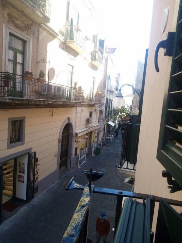 A narrow street with buildings on both sides, featuring balconies with potted plants, a visible storefront, and a pedestrian walkway.