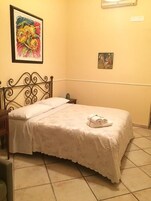 Una cama bien hecha con una colcha blanca y una toalla enrollada encima, una mesita de noche con una lámpara, y un cuadro en la pared.