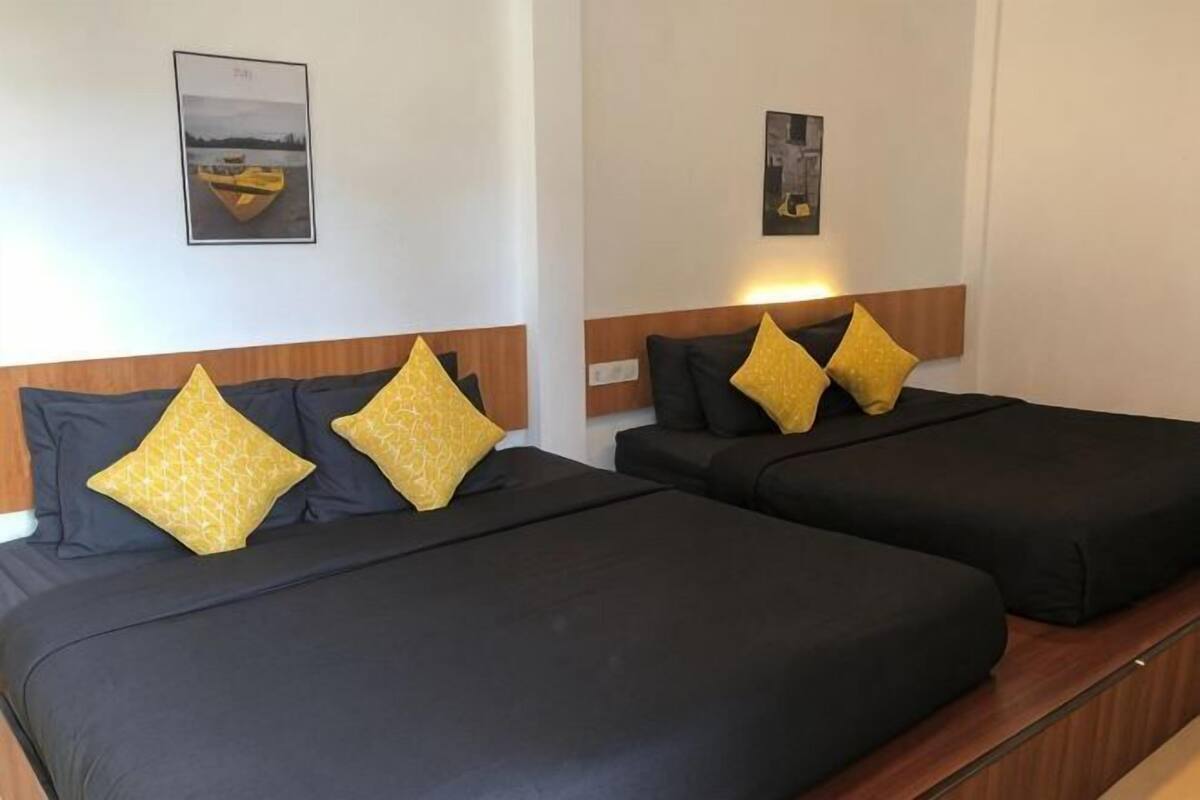 Two beds with black bedding and yellow pillows, a wooden headboard, and a framed picture on the wall.