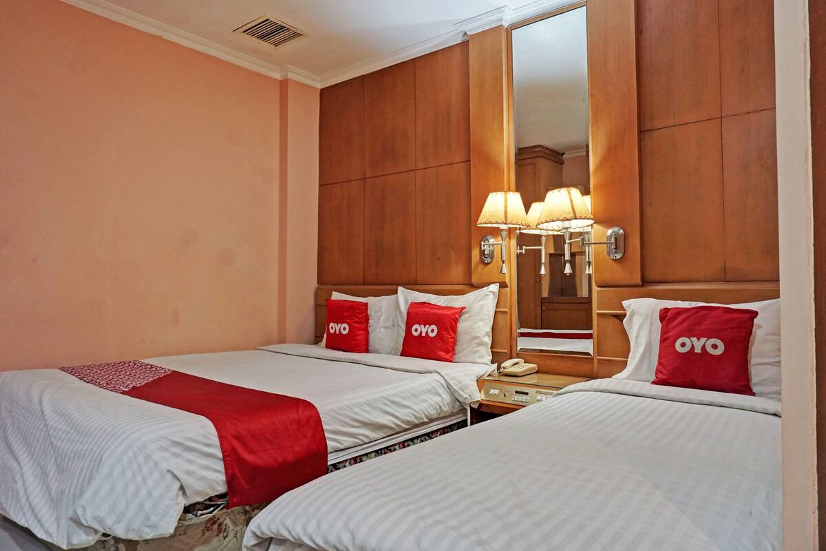 A hotel room with two beds, each with a red OYO branded pillow, a bedside table with a lamp, and a large mirror on the wall.