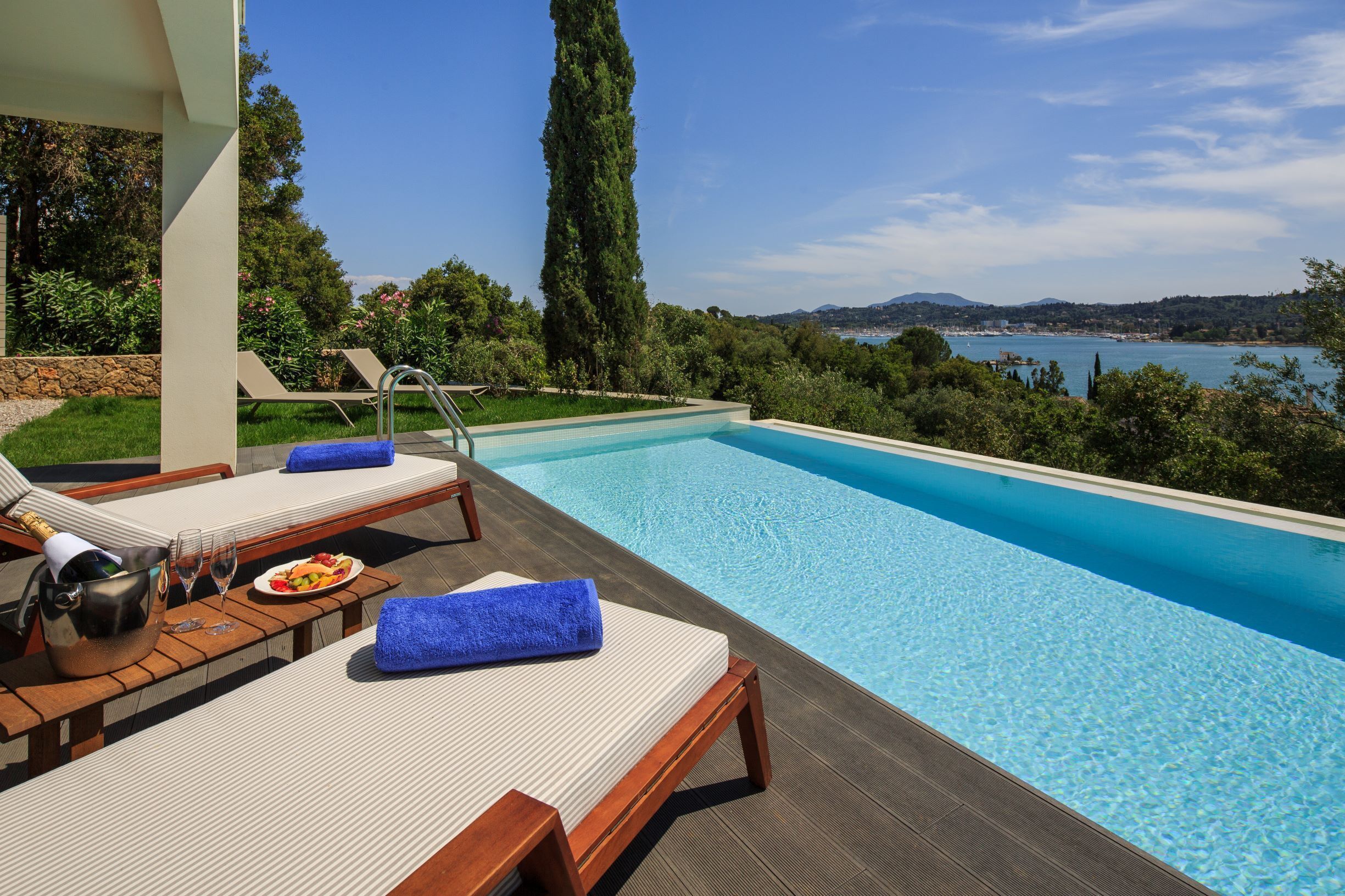 A poolside area with lounge chairs, a table with a bottle of champagne, and a view of the sea and hills.