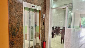Un ascensor con pantalla digital, un extintor y una puerta de vidrio que da a un comedor con mesas y sillas.