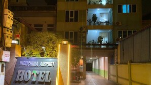 La entrada al Hotel Indochina Airport con un cartel, un camino que lleva a la entrada y un edificio con balcones y ventanas al fondo.