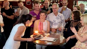 Un grupo de personas reunidas alrededor de una mesa con pastel y bebidas, sugiriendo un evento social o una celebración.
