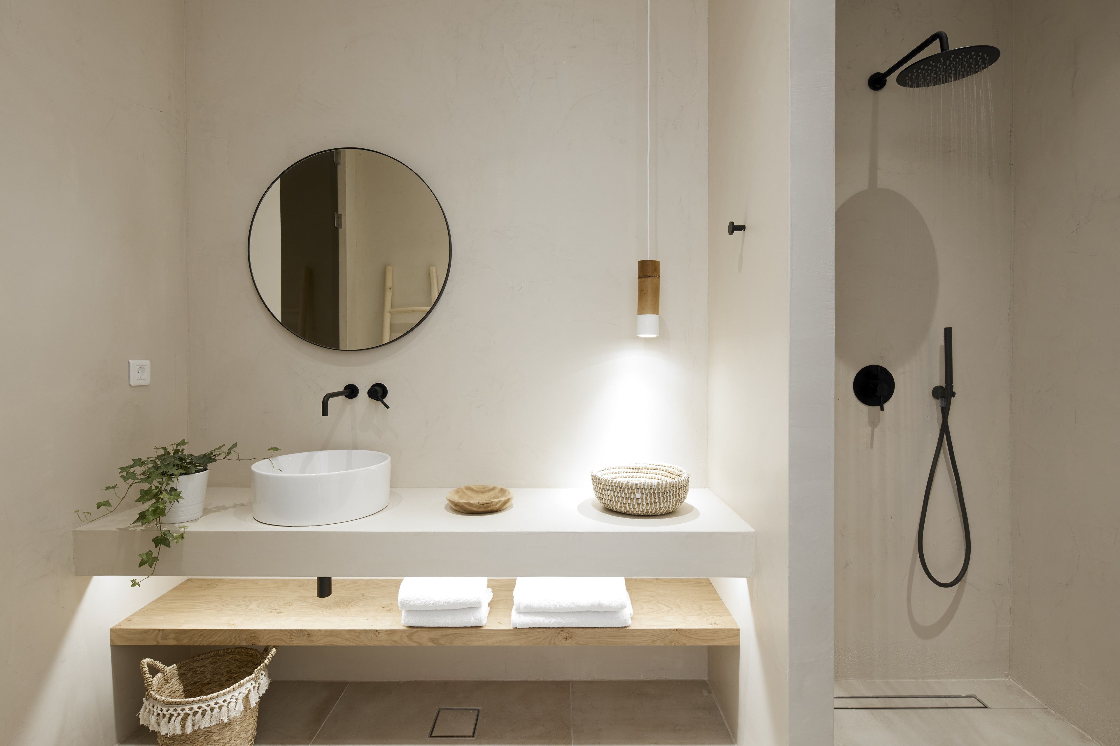 A modern bathroom with a round mirror, a white sink, a wooden vanity, a shower with a handheld showerhead, and a hanging light fixture.