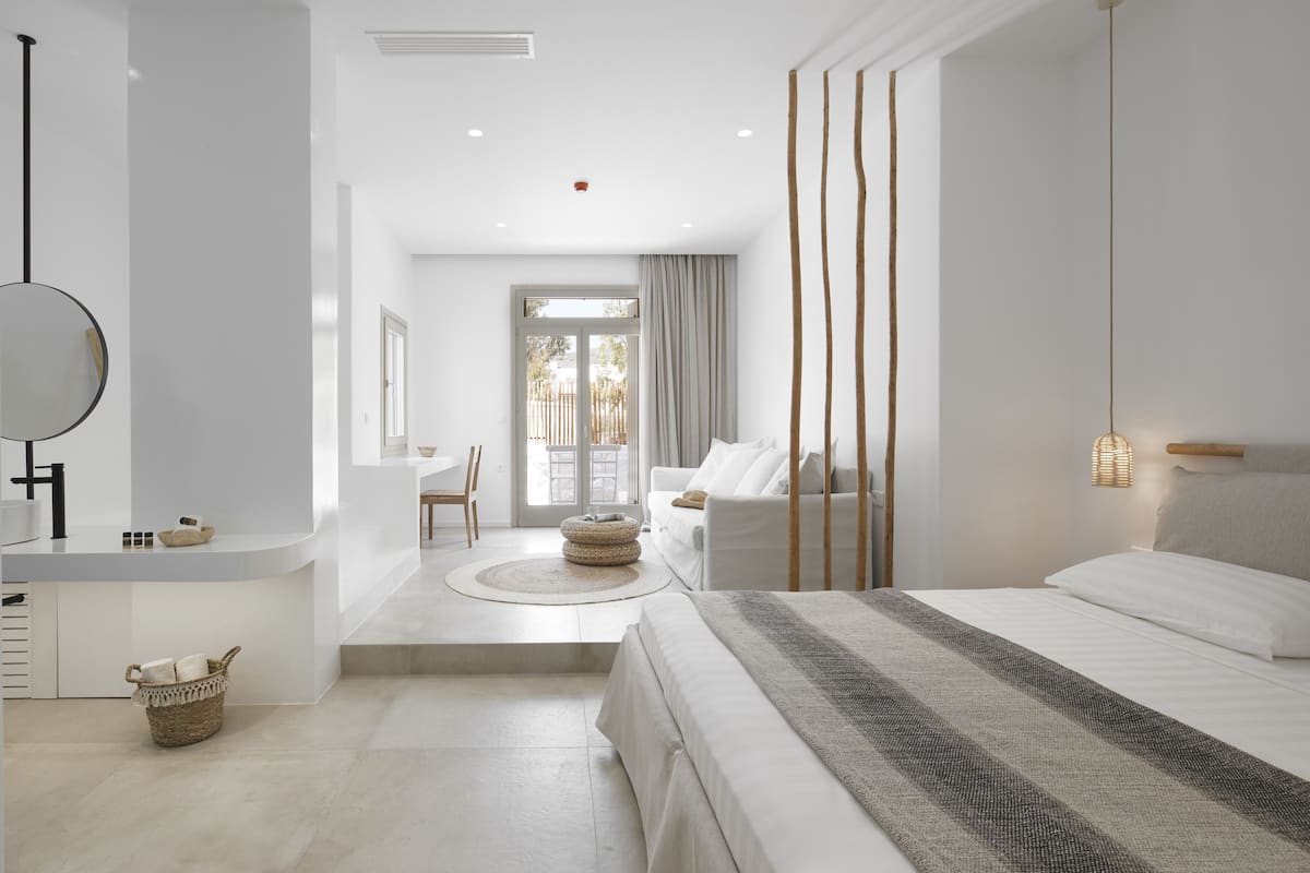 A modern hotel room with a large bed, a sitting area with a sofa and chairs, a small kitchen area, and a view of the outdoors.