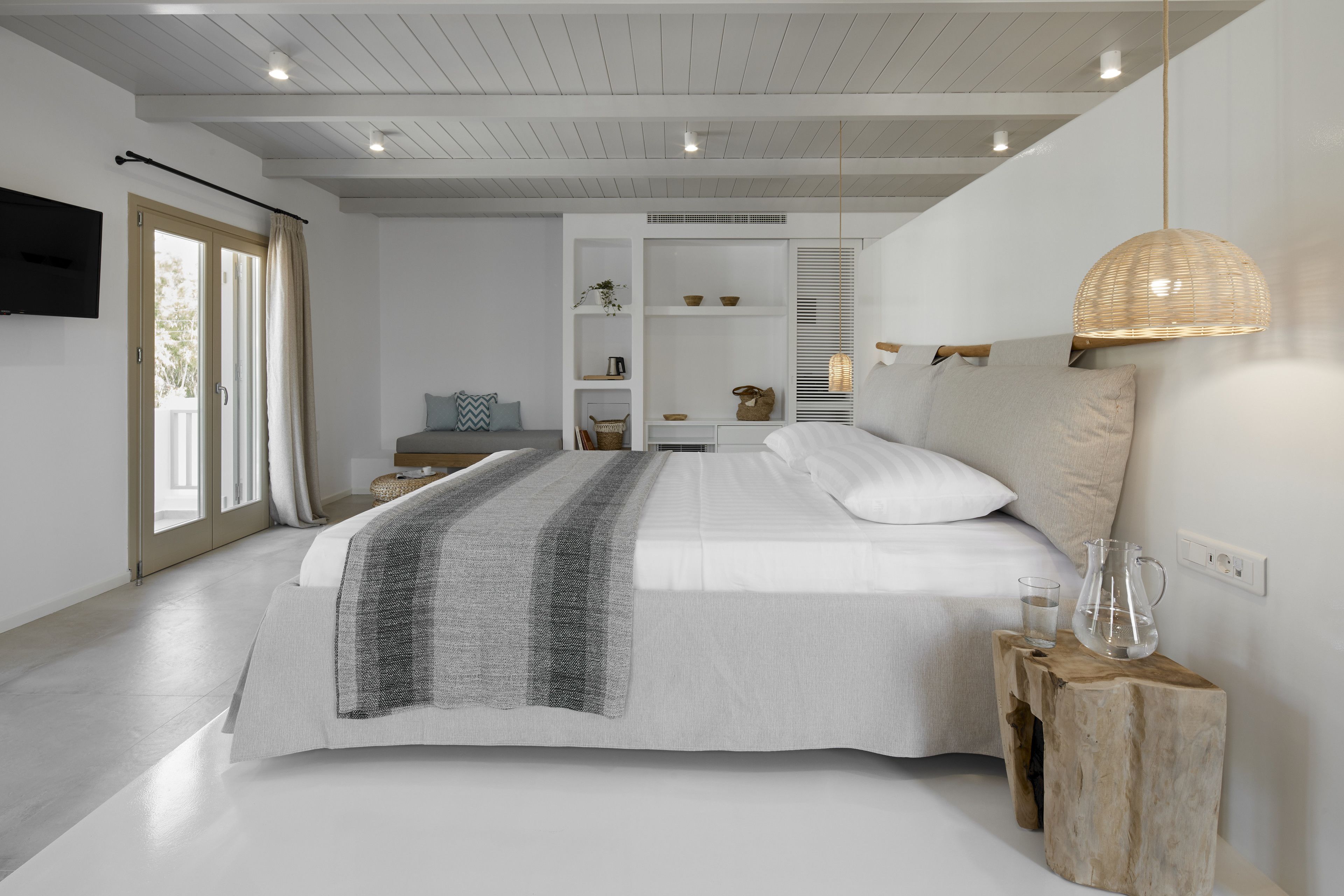 Un dormitorio moderno con una cama grande, una mesita de noche de madera, un televisor montado en la pared y un ventanal con cortinas.