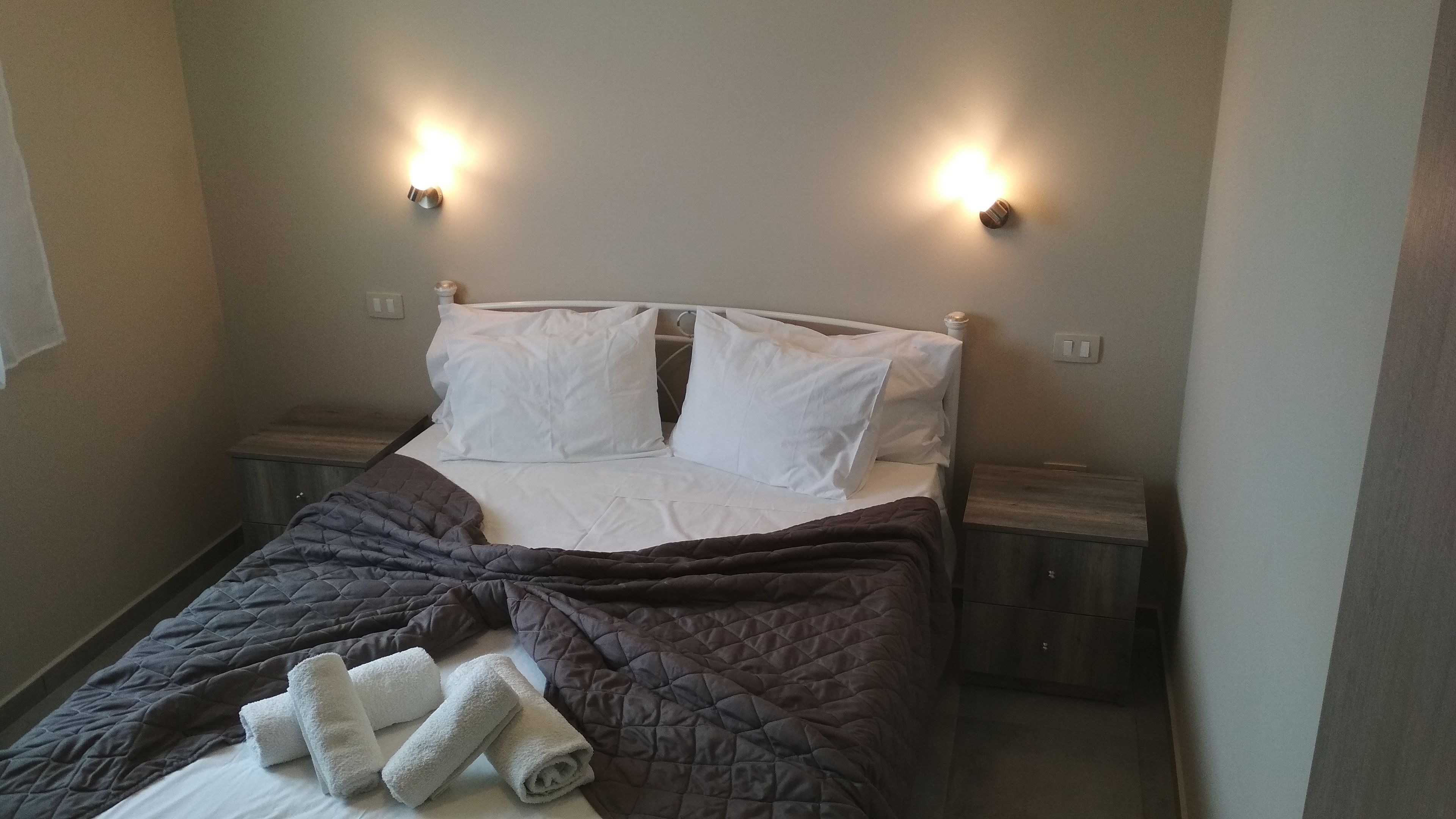 Ein Einzelbett mit dunkler Bettdecke, weißen Kissen und gerollten Handtüchern. Zwei Nachttische mit Lampen sind vorhanden.