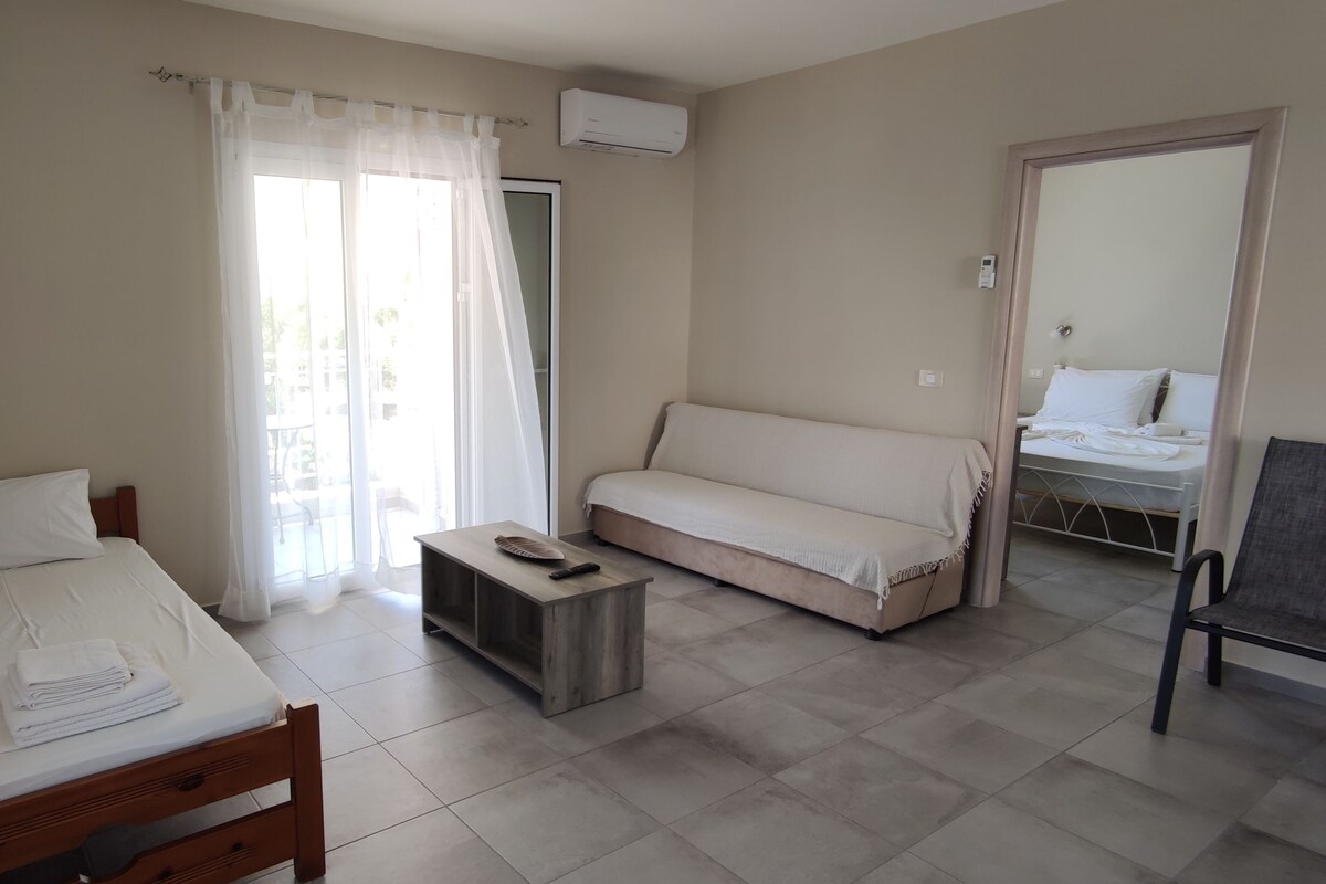 A hotel room with a bed, a sofa, a small table, and a chair. There is a window with curtains, an air conditioning unit, and a door leading to another room.