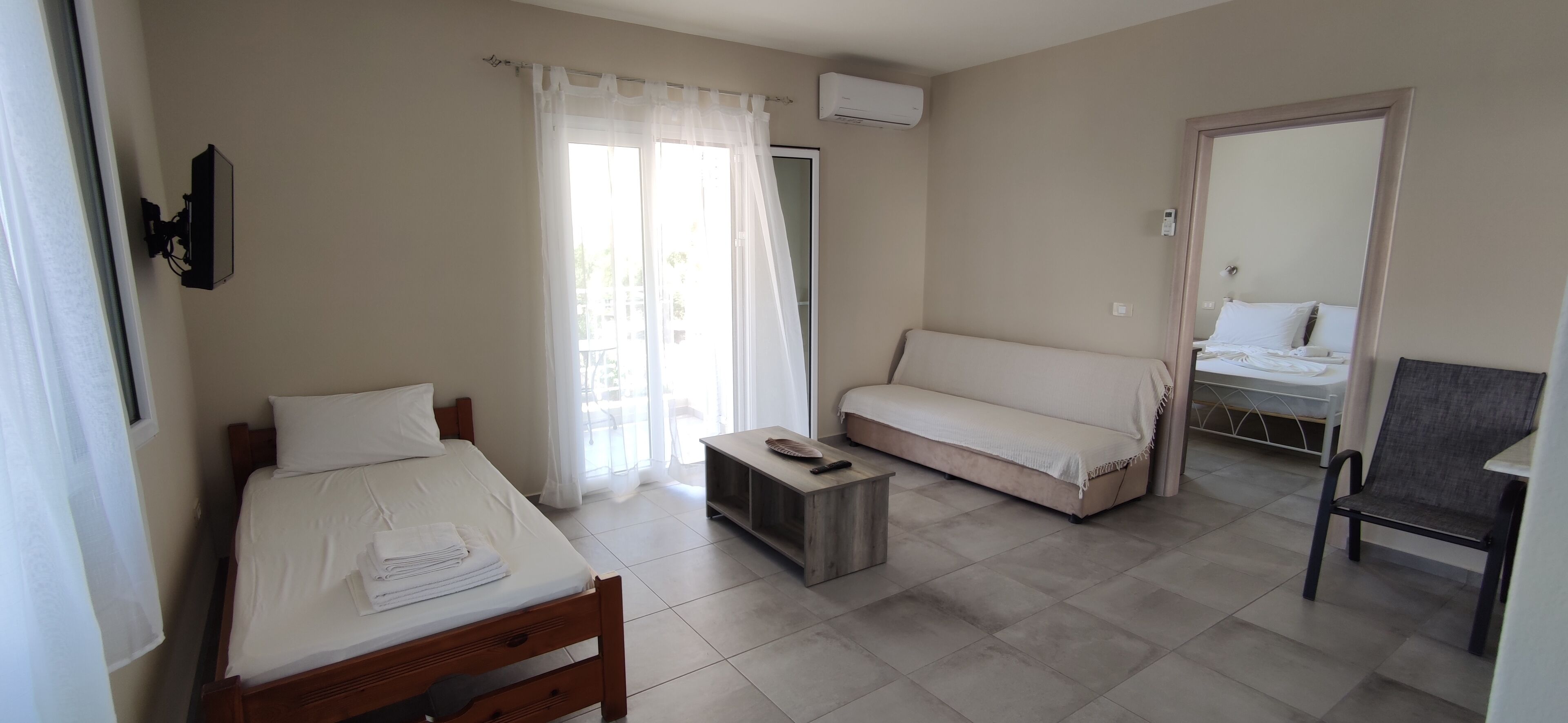 Ein Hotelzimmer mit einem Bett, einem Sofa, einem kleinen Tisch und einem Stuhl. Es gibt ein Fenster mit Vorhängen, eine Klimaanlage und eine Tür zu einem anderen Raum.