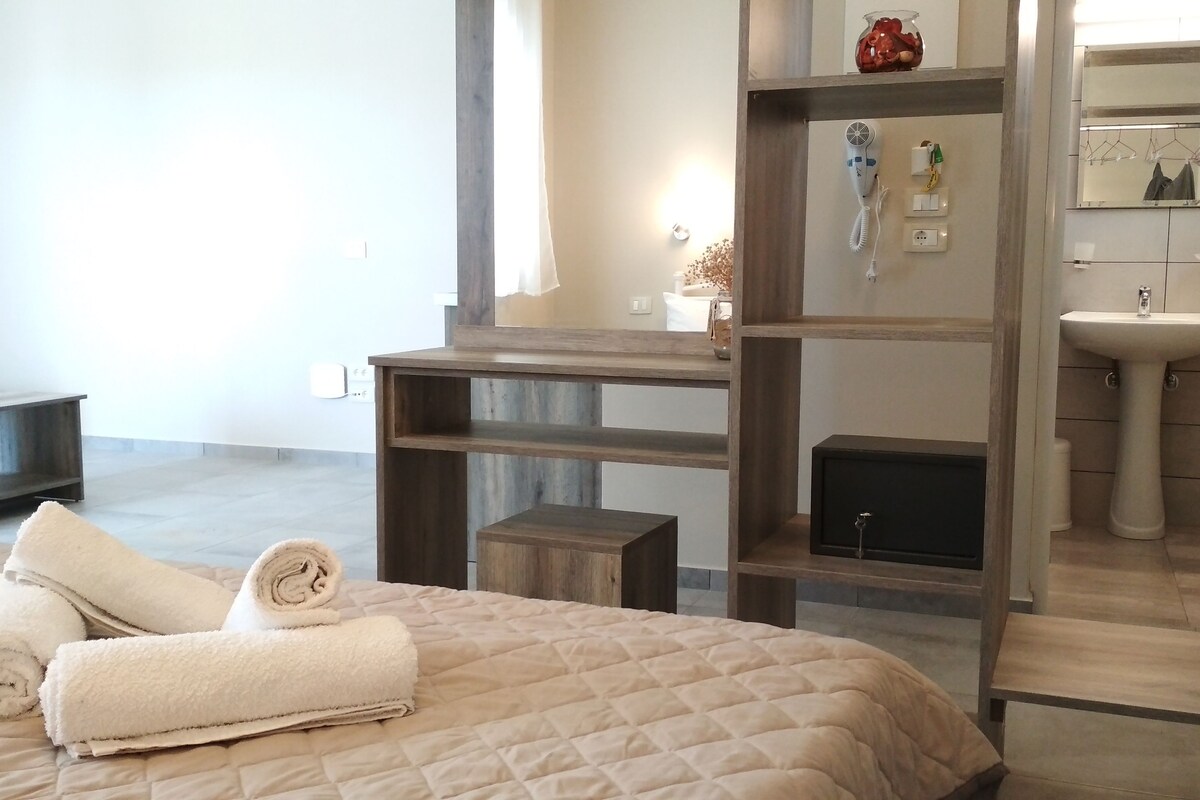 A hotel room with a bed, a wooden shelf unit, a bedside table, a mirror, a sink, and a towel on the bed.