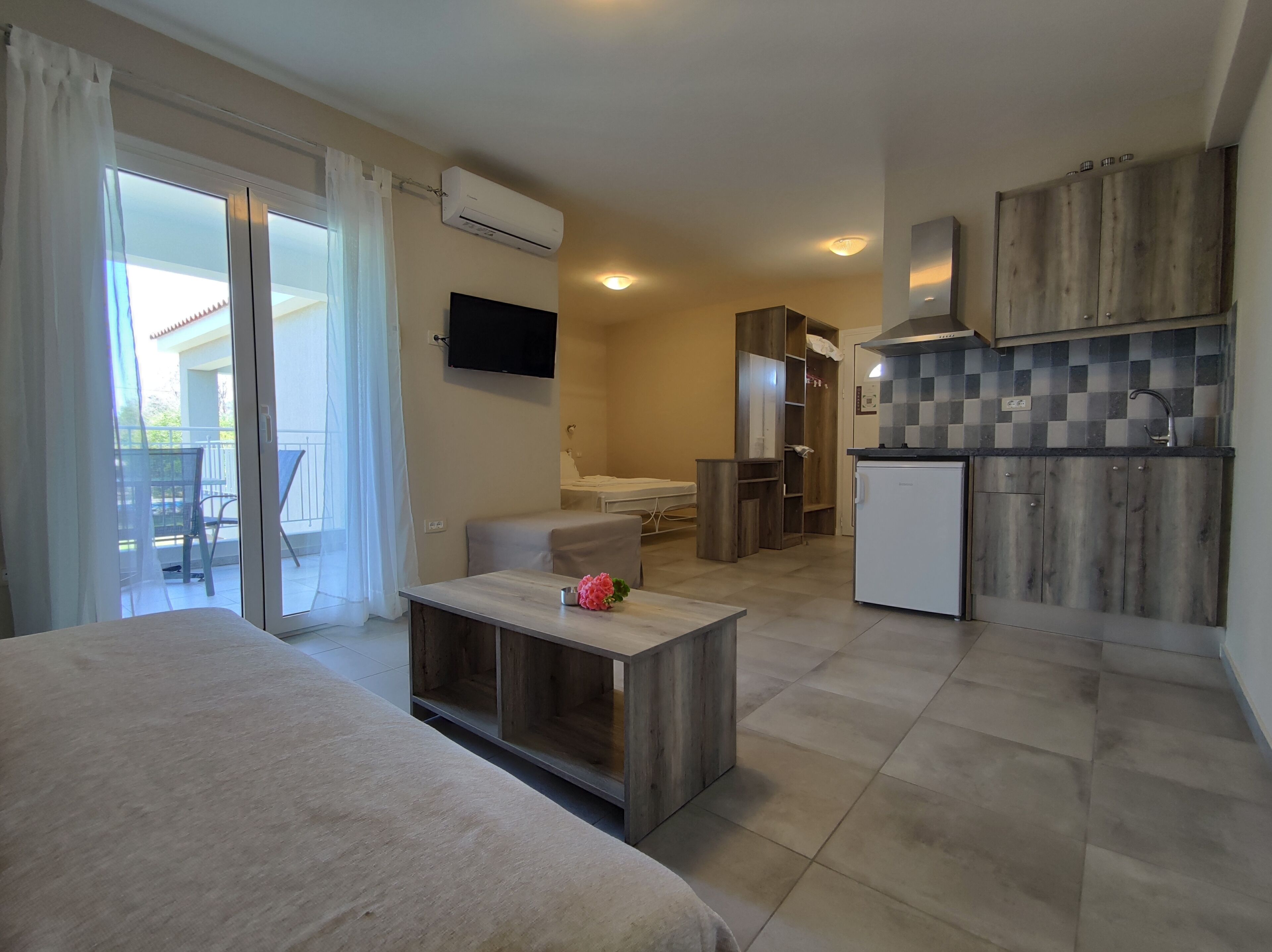 A modern hotel room with a bed, a small table with a flower, a TV, a kitchenette, and a sliding glass door to the balcony.