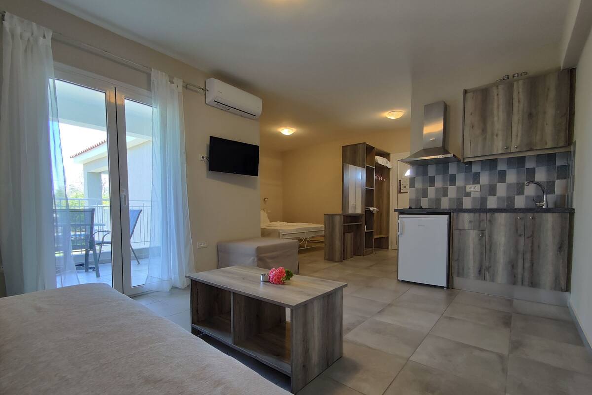 A modern hotel room with a balcony, a small kitchenette, a sofa, and a TV.