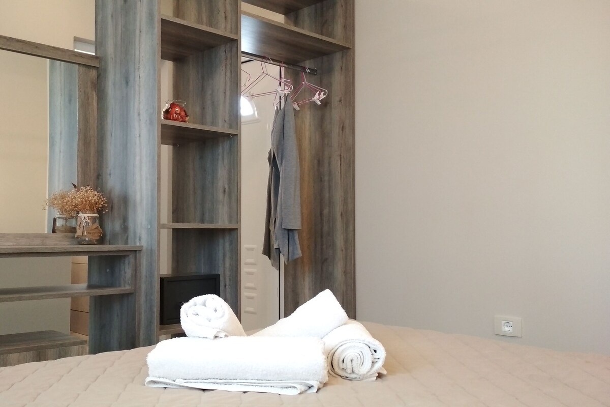 A bedroom with a large mirror, wooden shelving, a bed with rolled towels, and a hanging clothes rack.
