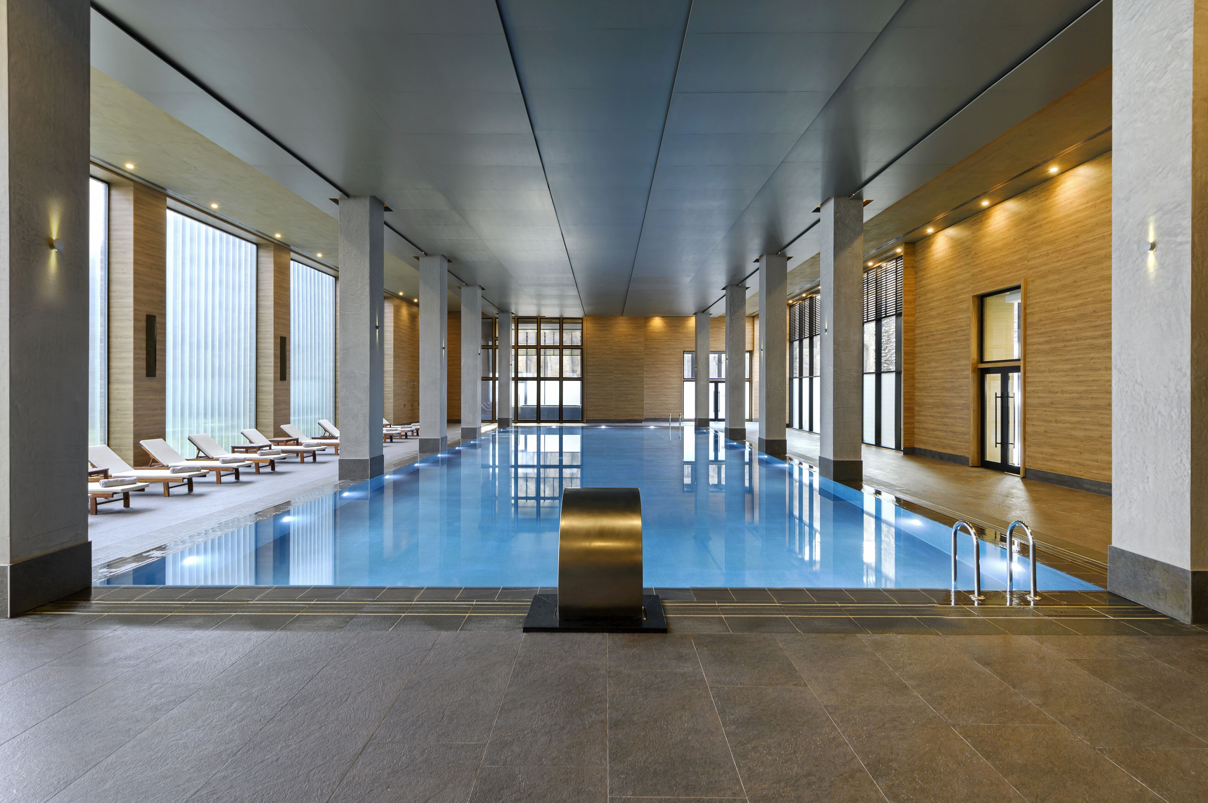 A large indoor swimming pool with clear glass walls, sun loungers along the sides, and a modern architectural design.