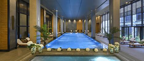 A large indoor swimming pool with a long, narrow shape, surrounded by lounge chairs and potted plants.