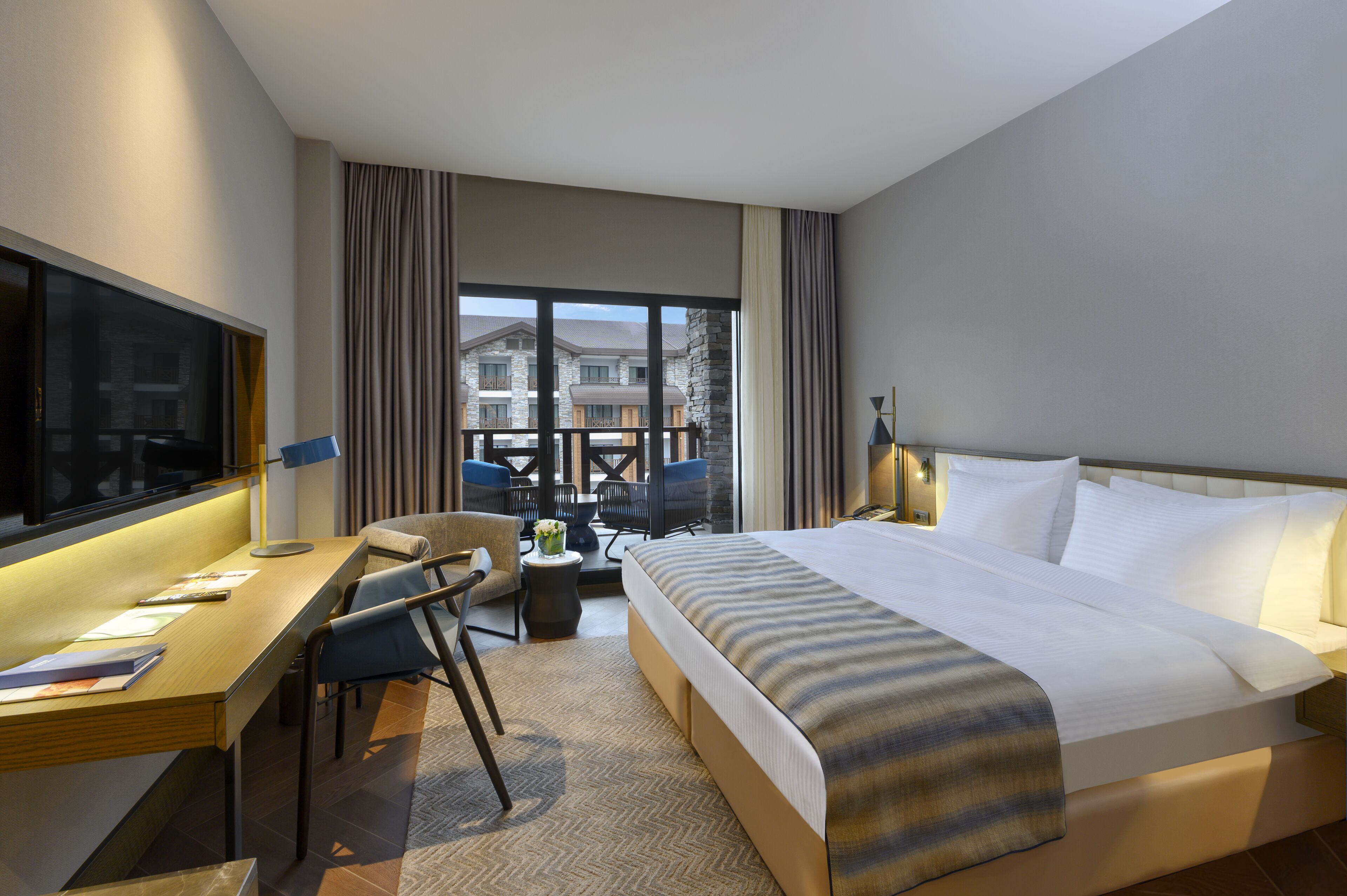 Habitación de hotel con una cama grande, un escritorio con una silla, un televisor y vistas a edificios a través de la ventana.