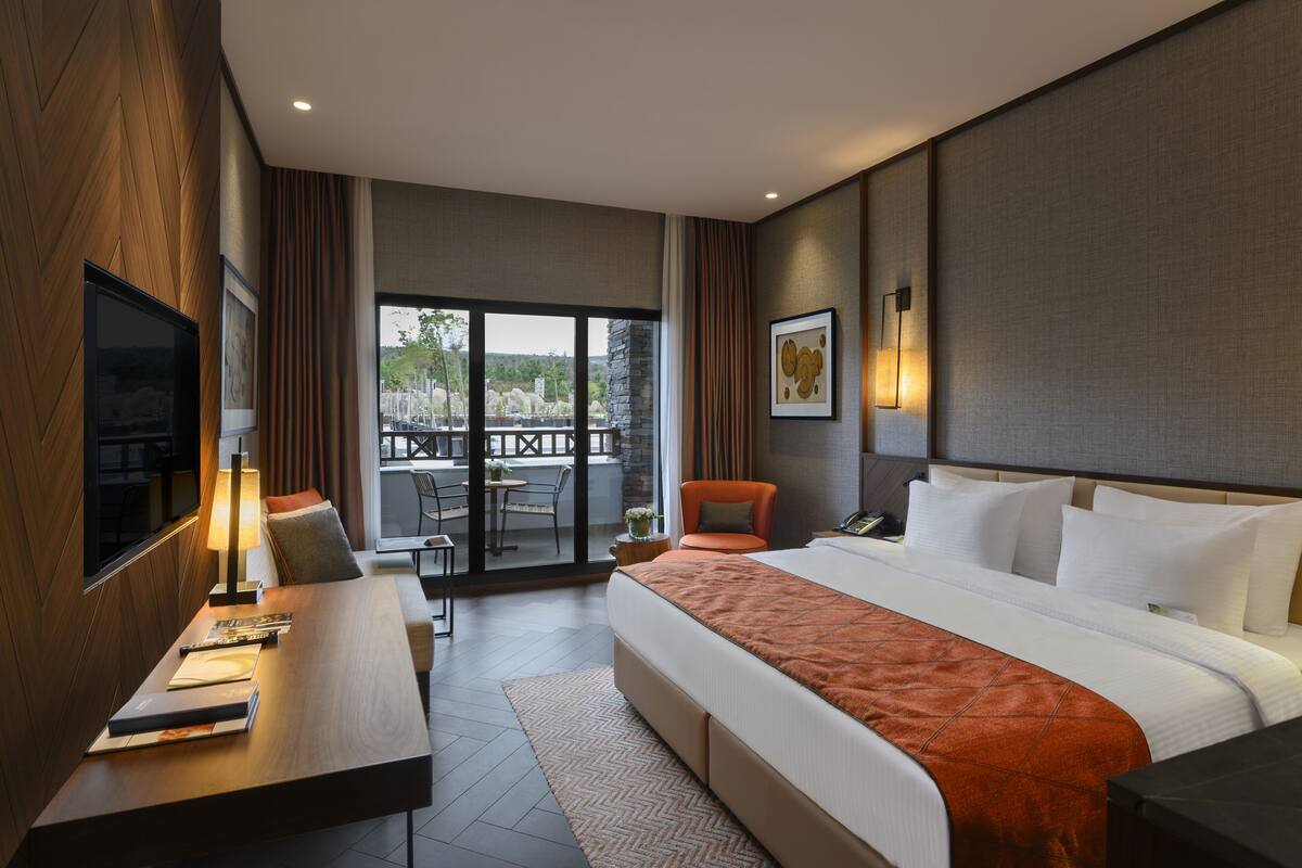Een hotelkamer met een groot bed, een bureau met lamp, een stoel, een televisie en een balkon met uitzicht op bomen.