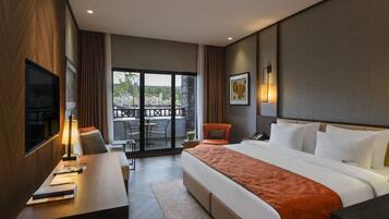 A hotel room with a large bed, a desk with a lamp, a chair, a TV, and a balcony with a view of trees.