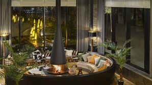 Un salon moderne doté d’une cheminée centrale, de sièges confortables et de grandes fenêtres offrant une vue sur l’extérieur.