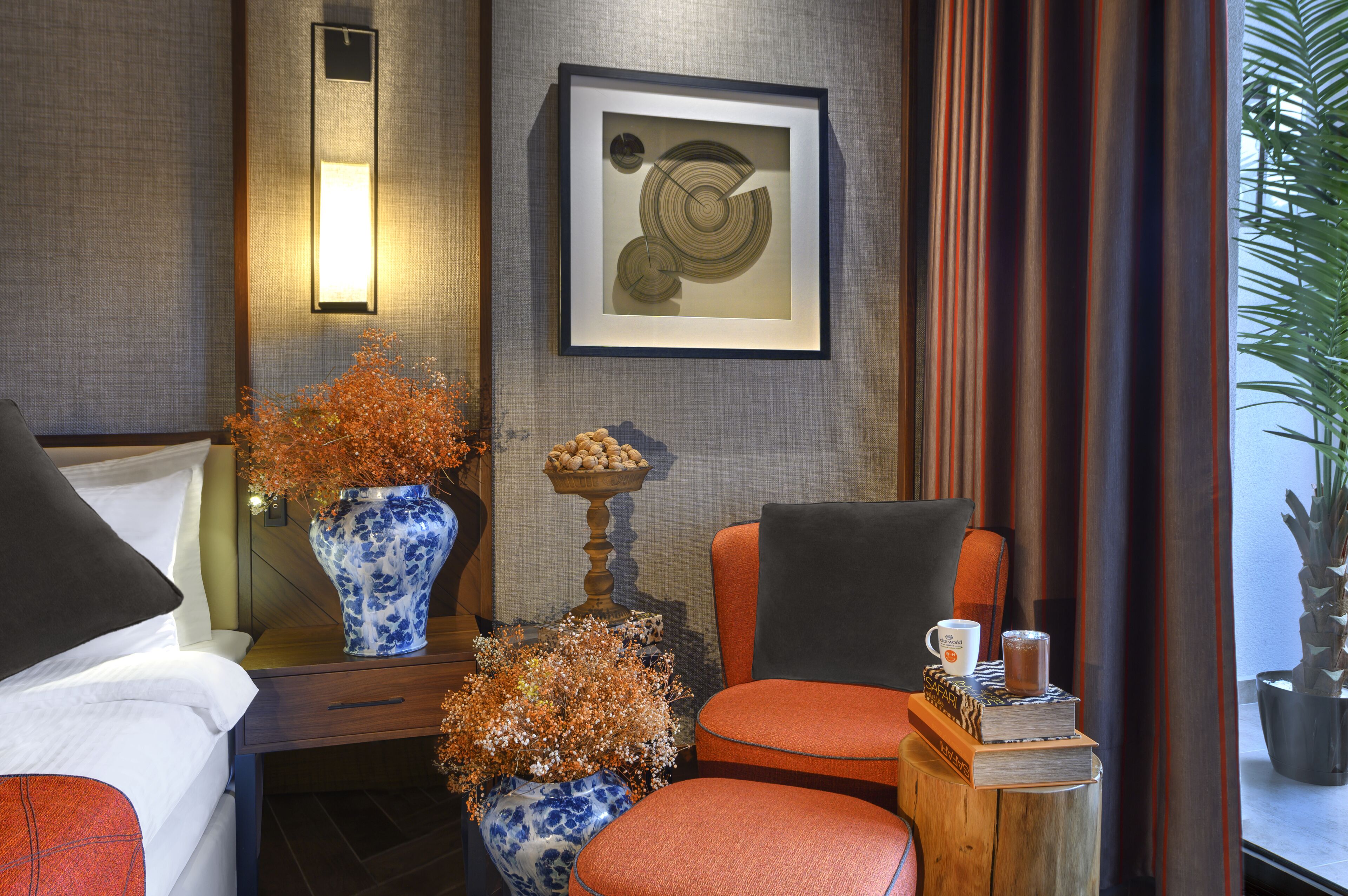 A hotel room with a bed, a chair, a small table with a vase, a framed artwork, and a potted plant.