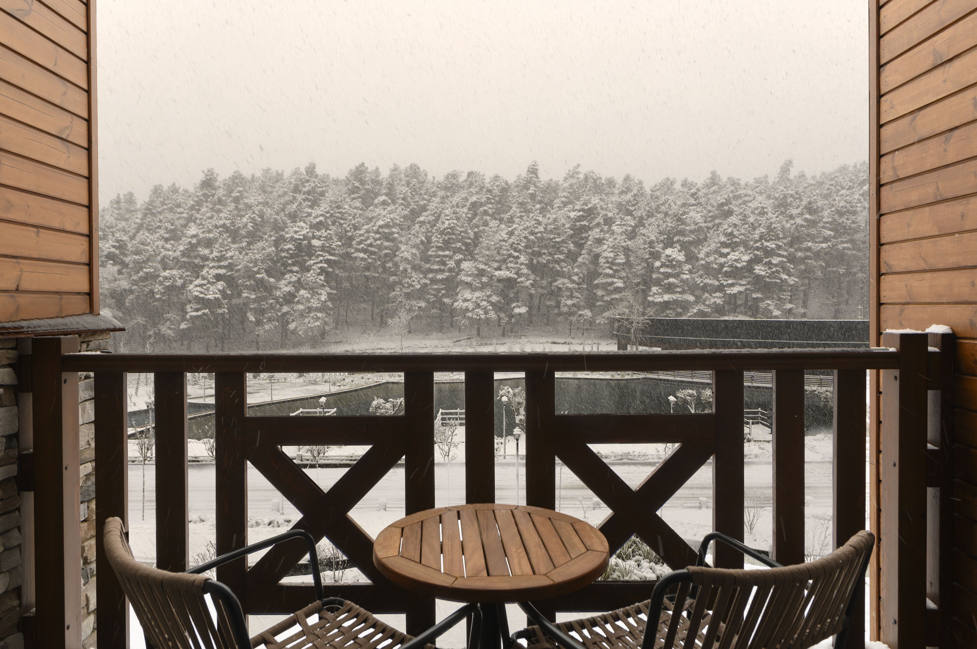 A wooden balcony with a table and chairs, overlooking a snowy landscape with trees.