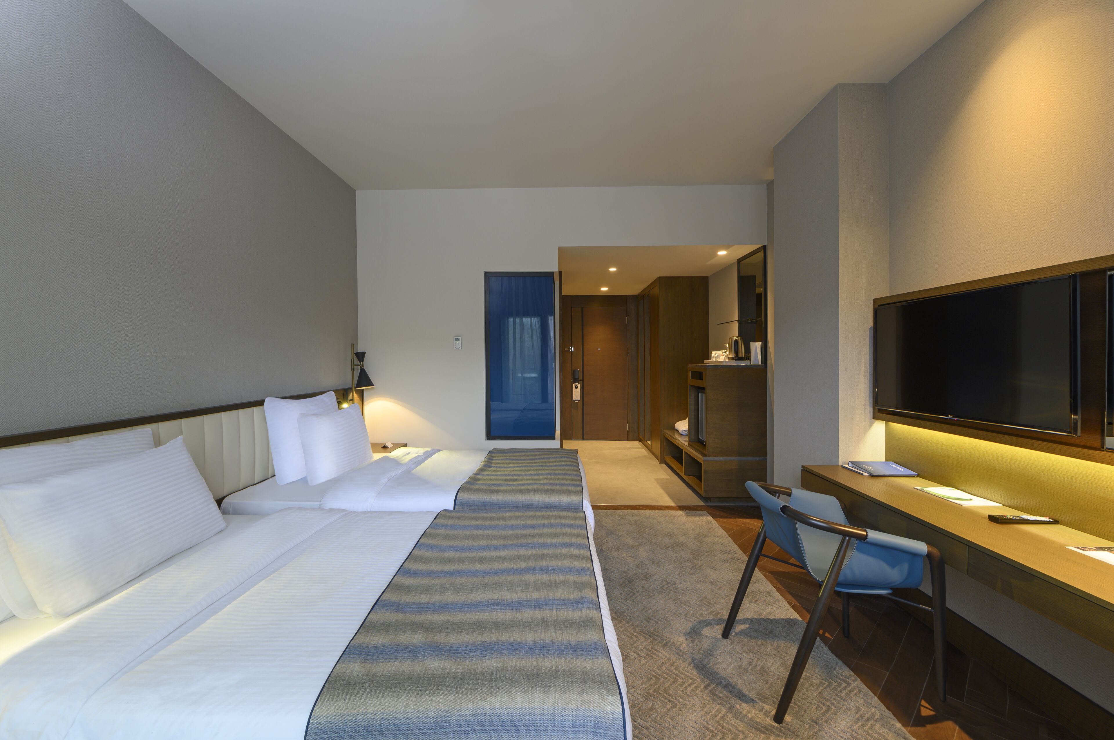 A hotel room with a bed, a desk, a chair, a TV, and a door leading to another room.