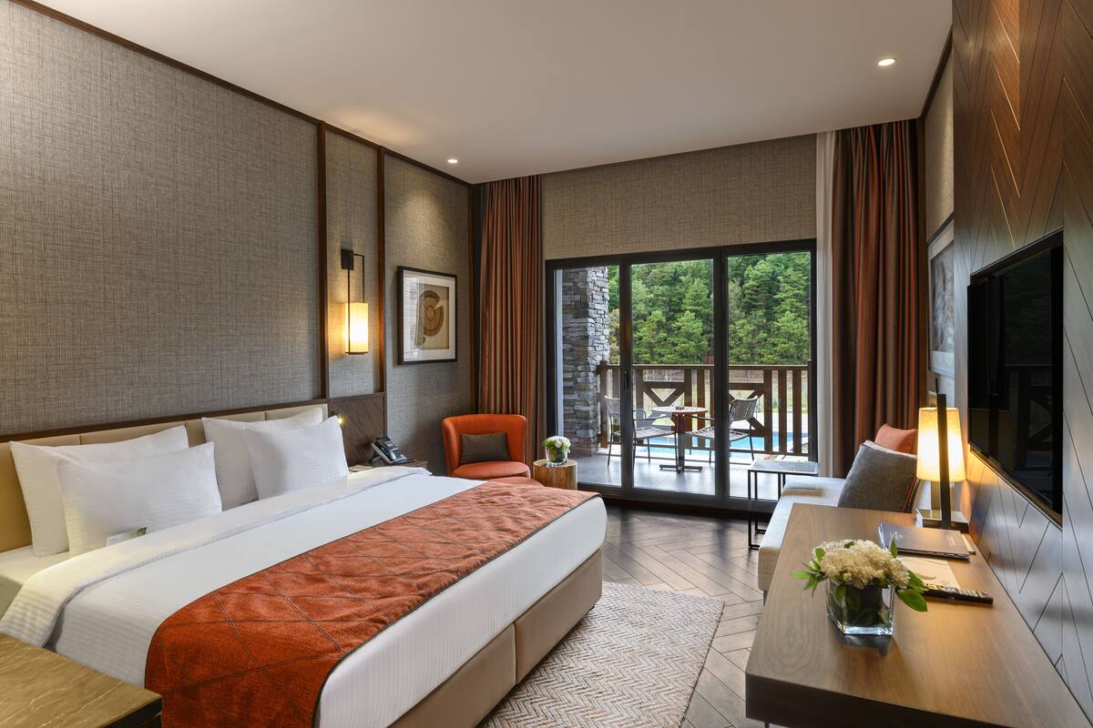 Een hotelkamer met een bed, een bureau met een lamp, een stoel, een klein tafeltje met bloemen, en uitzicht op een buitengebied met een zwembad.