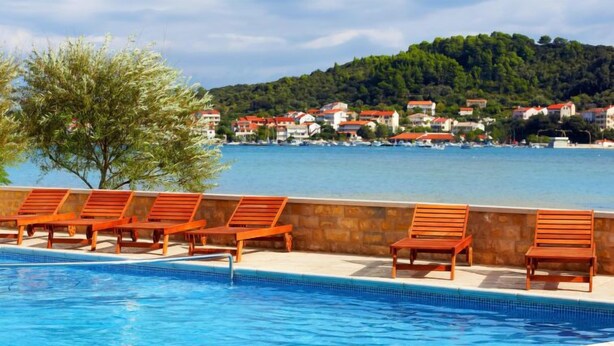 A swimming pool with wooden lounge chairs and a view of a coastal town with houses.