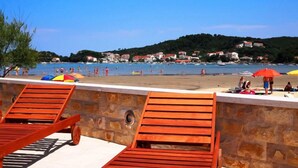 A beach with wooden lounge chairs and umbrellas, a stone wall, and a view of the sea and buildings in the background.